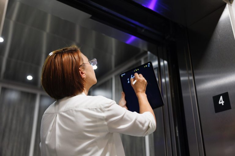 Zu sehen ist eine Prüfung der Aufzugssicherheit durch eine Prüferin mit Prüftableau und elektronischem Pen in der Hand. Ihr Blick ist nach oben gerichtet auf eine elektronische Anzeigeeinheit.